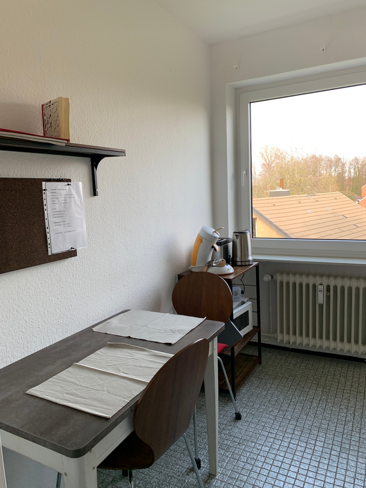 A small office space is featured, containing a desk and two chairs, with a shelf above for storage. A window allows natural light, overlooking rooftops. An electric kettle and microwave are on a side table, alongside a coffee machine. A radiator is positioned beneath the window.