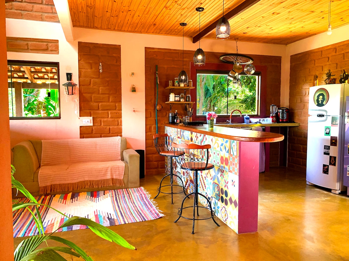 The open-concept living space features a colorful kitchen island with bar stools, adjacent to a cozy seating area. Natural light filters through large windows, highlighting the warm tones of the walls and wooden ceiling. Decorative elements add character to the inviting atmosphere.
