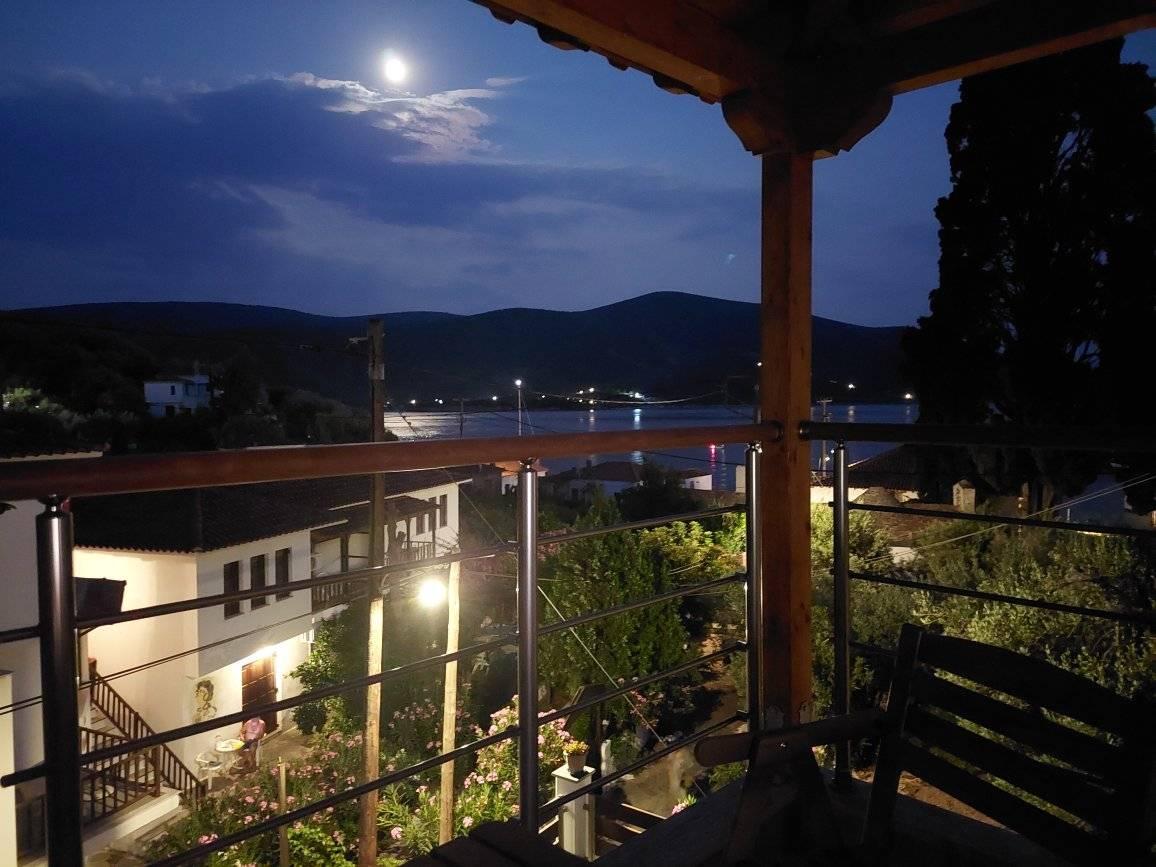 An evening view from a balcony captures the tranquil scenery under a bright moon. The outlines of distant hills are visible, along with lights from houses reflecting on the water, creating a serene atmosphere.
