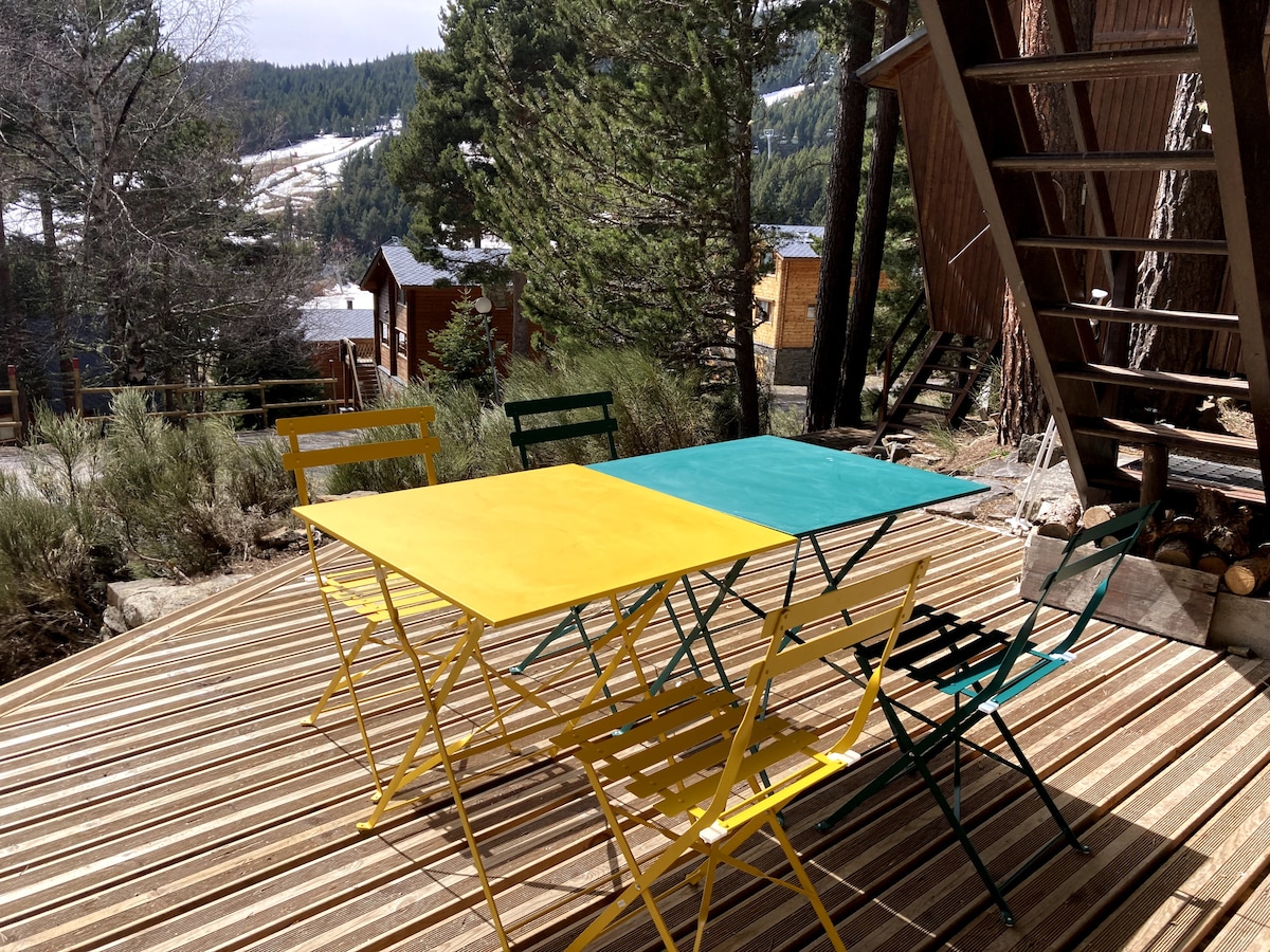 An outdoor terrace features a brightly colored table with yellow and teal surfaces, surrounded by folding chairs in various shades of green and yellow. The setting is framed by tall trees, with a view of distant mountains visible in the background.
