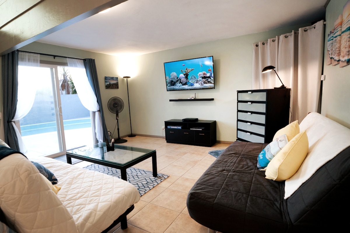 A welcoming living area is designed with a neutral color palette. A comfortable sofa bed is paired with a matching lounge chair. A large glass door allows natural light, revealing a view of the pool area. A 75" TV is mounted on the wall, enhancing entertainment options.