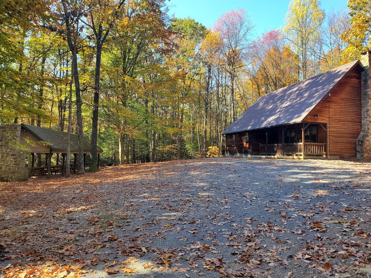 A spacious cabin is set amidst a forest of trees displaying vibrant autumn colors. A gravel area is visible in front of the cabin, with fallen leaves scattered on the ground. An outdoor pavilion is located nearby, enhancing the natural setting.