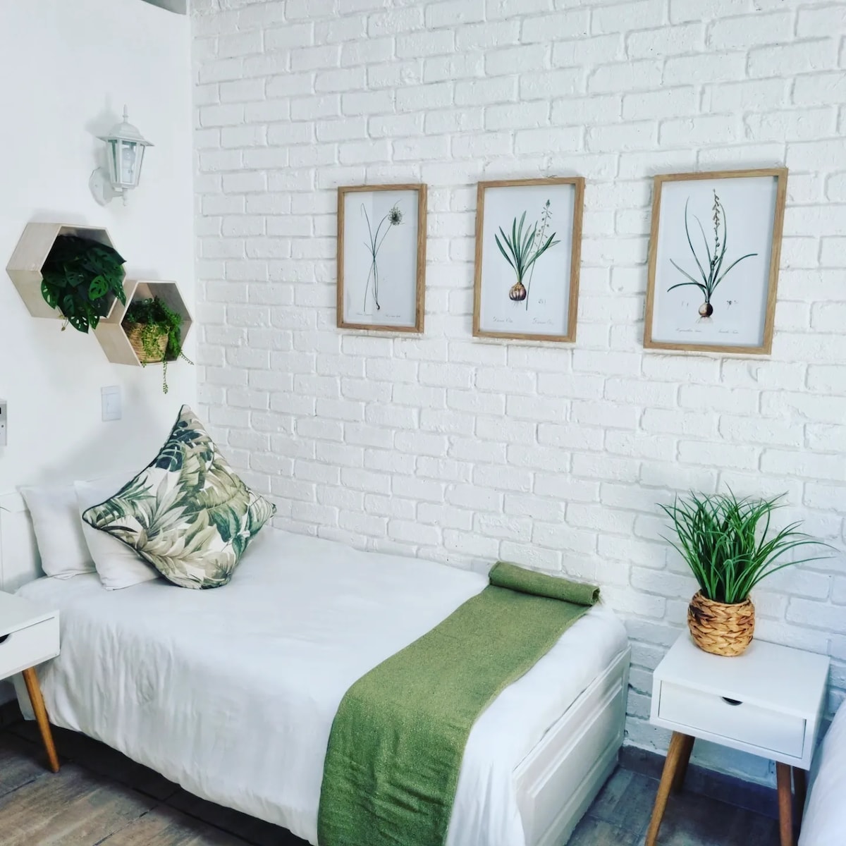 A bright room features a single bed with a white duvet and a decorative green throw. Framed botanical prints adorn the white brick wall, while a small side table with a plant sits beside the bed. A wall-mounted light fixture adds additional illumination.