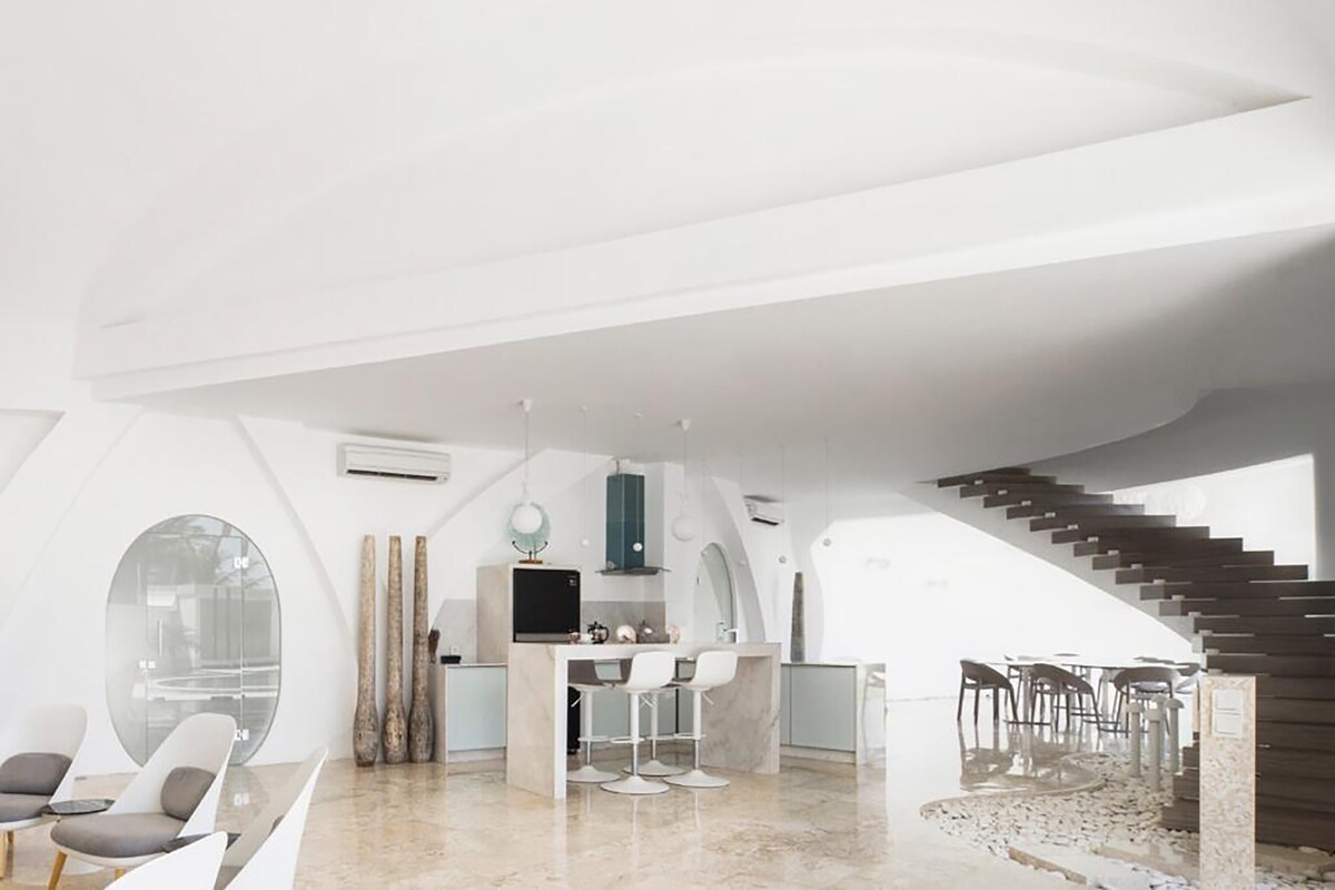 An open-plan living area showcases a modern kitchen with sleek appliances and a spacious dining table. Curved white walls and large circular windows enhance natural light. Elegant seating arrangements consist of minimalist chairs, and a gracefully designed staircase leads to the upper level.