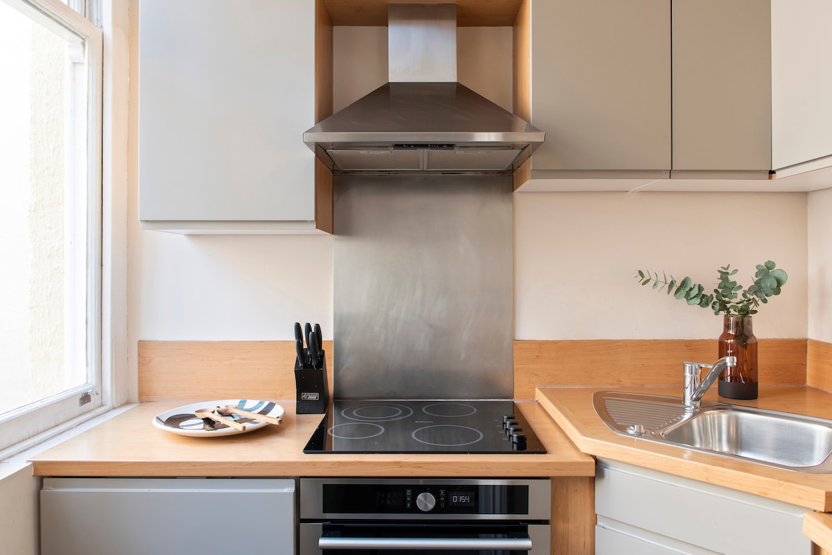 A modern kitchen area features a stainless steel extractor hood above a smooth, four-burner electric hob. A wooden countertop runs along the kitchen, complemented by sleek cabinetry. A sink is positioned nearby, and a small decorative plant adds a touch of greenery.