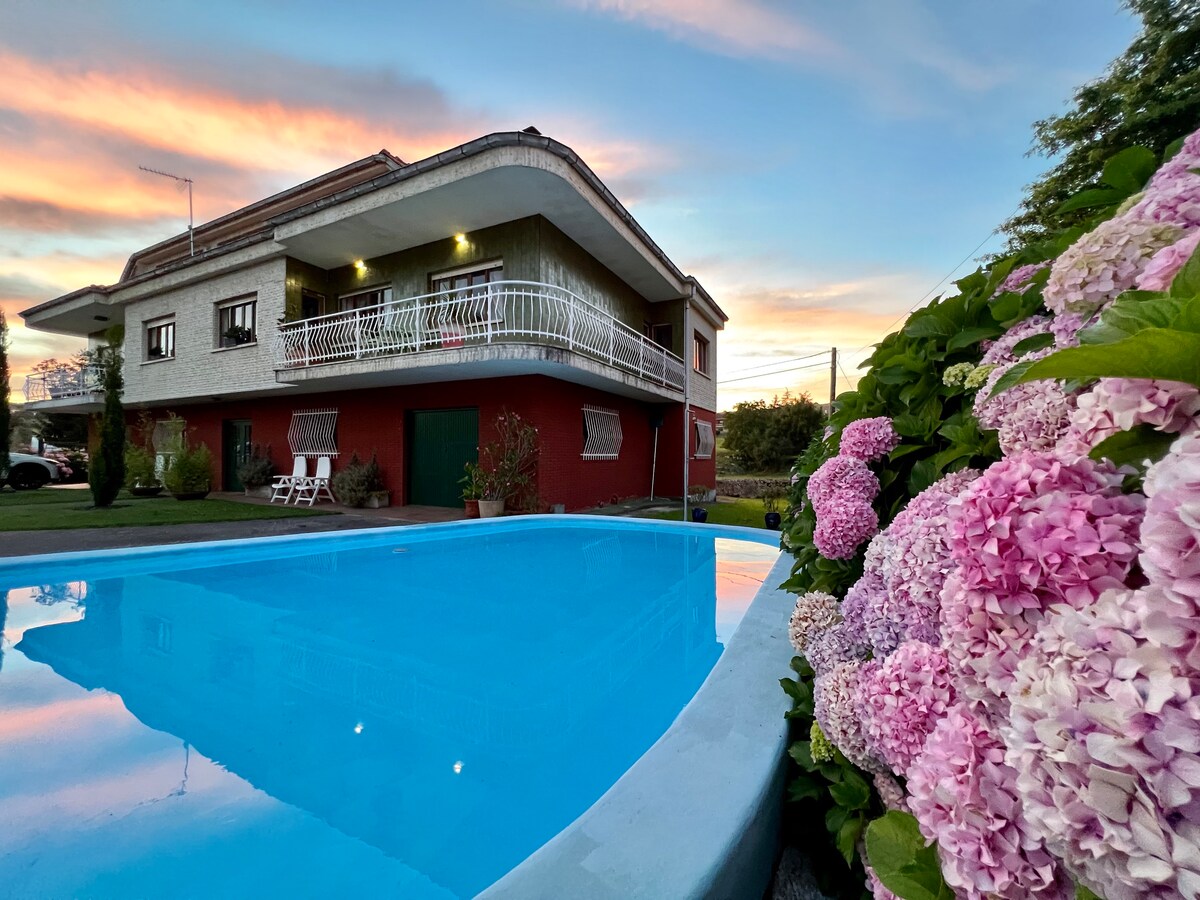 The image features a spacious two-story house with a balcony, nestled among lush greenery. A clear blue pool reflects the colorful sunset, while vibrant pink and white flowers border the poolside, adding a fresh, natural element to the scene.