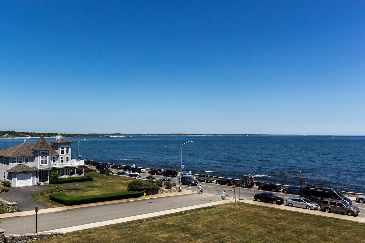 A view of the coastline reveals the Atlantic Ocean under a clear blue sky. The shoreline is lined with lush greenery and a parking area for nearby visitors. Cars are parked along the road, providing access to the waterfront.