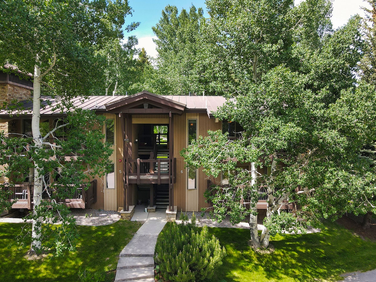 The exterior of the condo showcases a two-story structure surrounded by lush greenery and mature trees. A well-maintained pathway leads to the entrance, with natural grass and landscaped areas enhancing the tranquil outdoor setting.