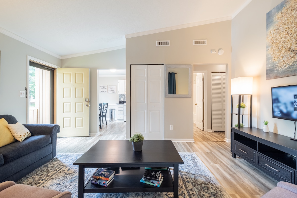 The living area features a dark wood coffee table positioned centrally, complemented by a dark sofa with yellow and white accent pillows. A large television is placed on the media console against the wall. Natural light floods in through a doorway leading to a kitchen and another room.