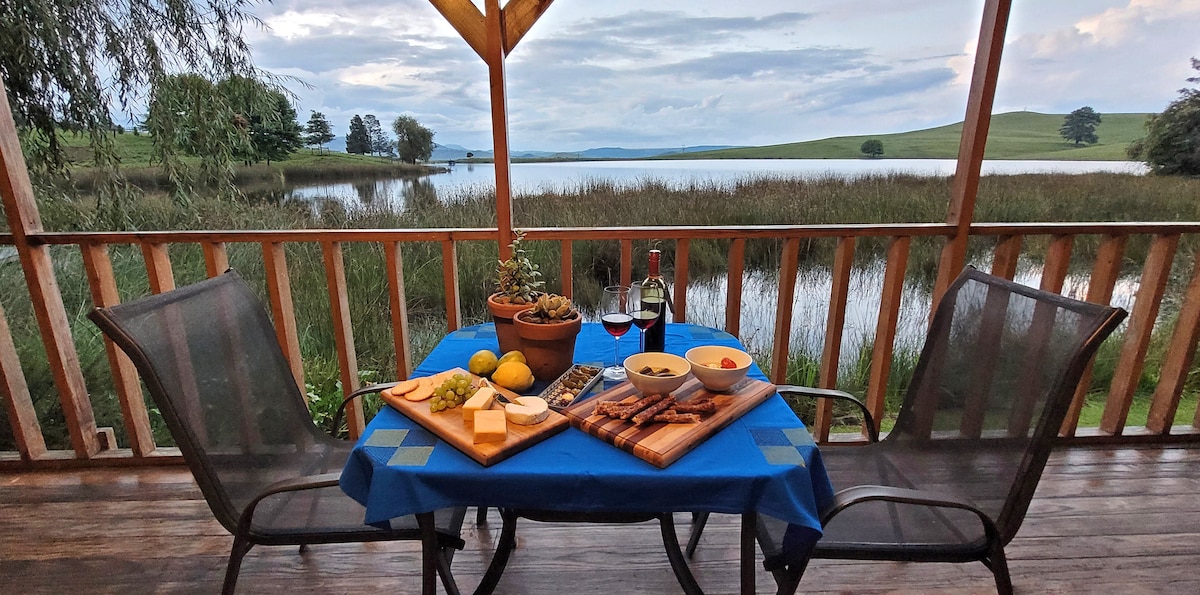 A dining setup is presented on a wooden verandah, featuring a blue tablecloth with a selection of cheeses, crackers, and fruits. Two chairs frame the table, while a bottle of red wine and glasses are visible. A tranquil dam and lush greenery serve as a picturesque backdrop.