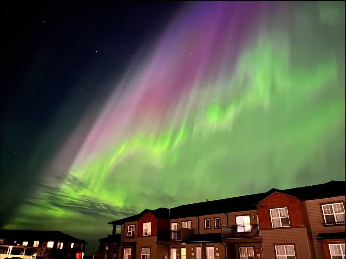 A vibrant display of the Northern Lights fills the evening sky, with swirling hues of green and purple. Below, modern townhouses with balconies are partially illuminated, creating a contrast against the natural spectacle above.
