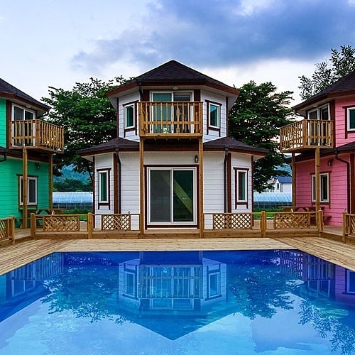 A modern vacation home is centered around a clear blue pool, flanked by colorful two-story houses in green and pink. Each house features balconies, and expansive glass doors provide views of the pool area and surrounding greenery.