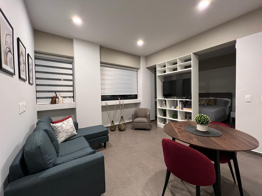 A modern living space features a comfortable seating area with a dark blue sofa and an armchair. A round dining table with red chairs sits adjacent to a shelving unit housing a television and decorative items. Natural light filters in through large windows covered with light blinds.