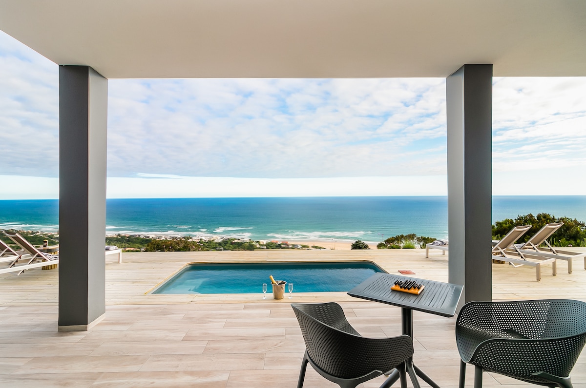 An outdoor terrace area is shown featuring a private pool with a clear view of the ocean beyond. Two modern black chairs surround a small table, which holds a snack. Sun loungers are positioned in the background, and the sky displays varying shades of blue and white.