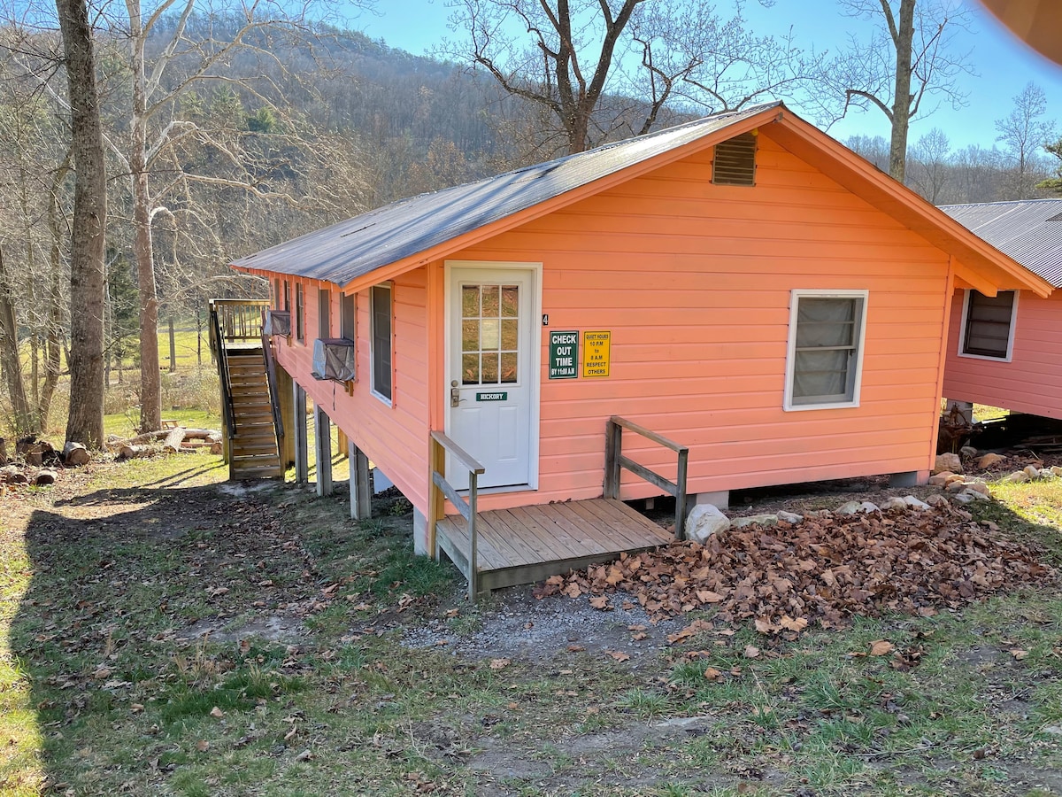 A cabin with a bright orange exterior is set amongst trees and rolling hills. The entrance features a wooden deck leading to the door, marked with signage. Steps ascend to an upper level, inviting guests to explore the surrounding landscape.