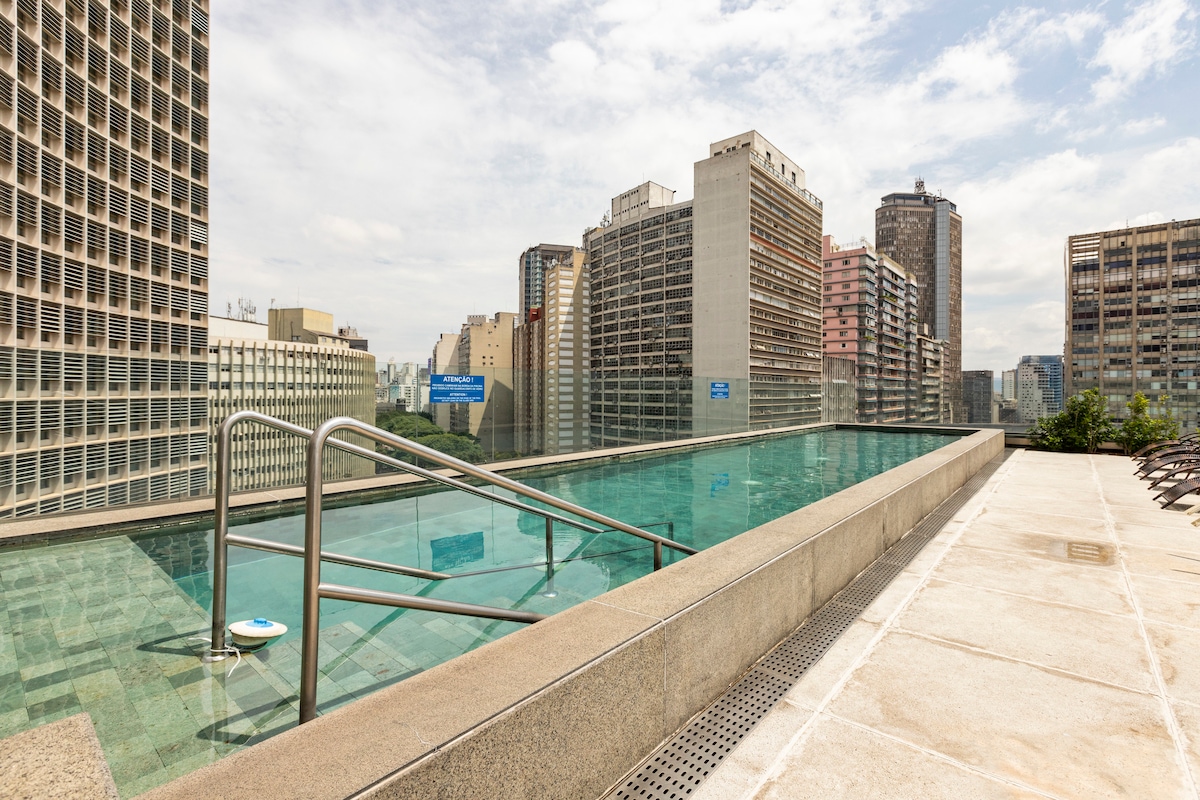 An infinity pool is depicted, offering a view of the surrounding city skyline. The water's edge blends with the horizon, and handrails are visible for safety. Lounge chairs are arranged along the pool deck, inviting relaxation. The clear sky and tall buildings create an open atmosphere.