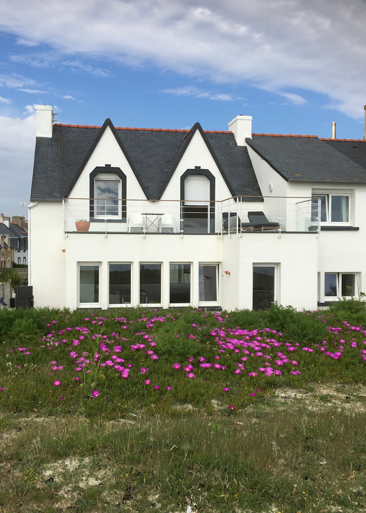 The exterior of a charming white house features a sloped roof and large windows, allowing natural light to fill the interior. A balcony extends from the upper level, offering views of the landscape. Vibrant pink flowers bloom in the foreground, enhancing the serene setting.