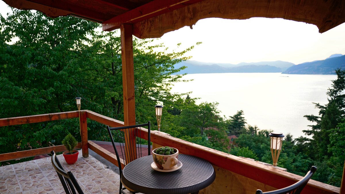 A wooden terrace overlooks a serene body of water surrounded by lush greenery. A round table with two chairs is set, featuring a small planter and a bowl of fresh fruit. Soft lighting from lanterns enhances the tranquil ambiance.