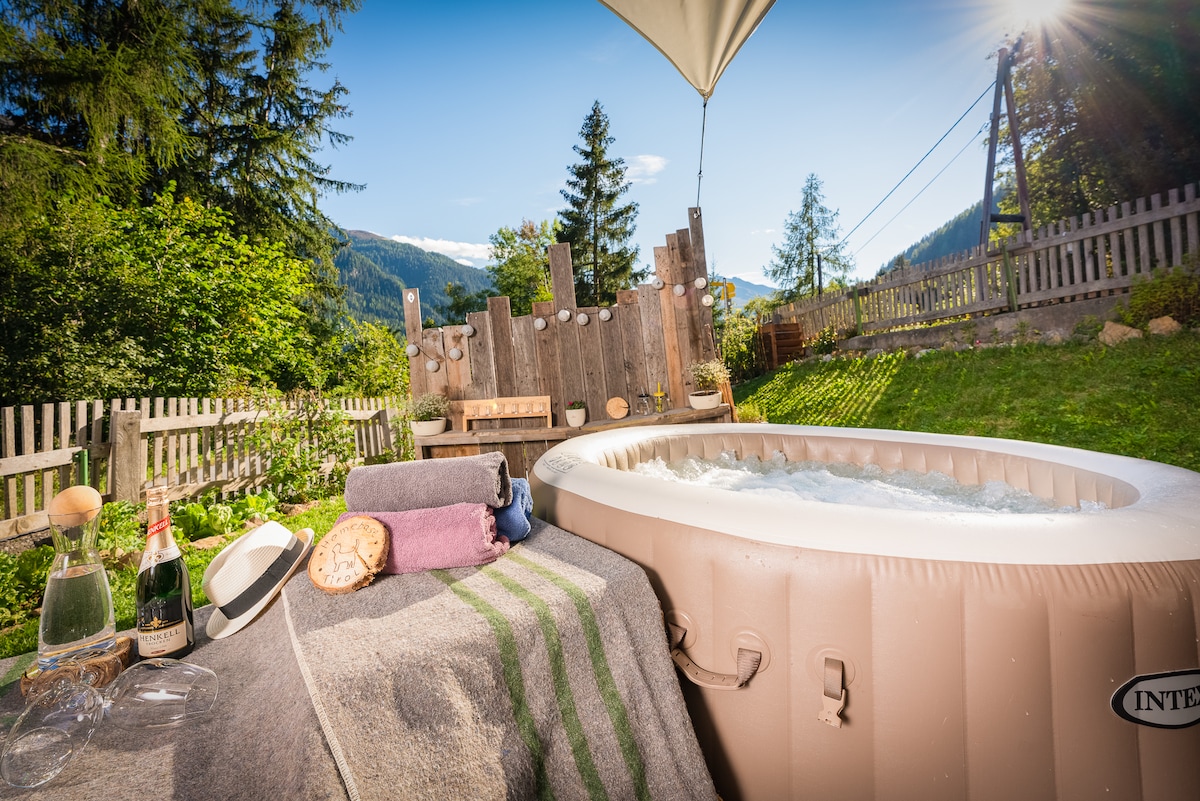 An outdoor space features a private hot tub surrounded by greenery and wooden fencing. A picnic table and benches are visible in the background, with mountains framed by trees. Towels and a bottle of champagne are placed on a cozy blanket beside the tub.