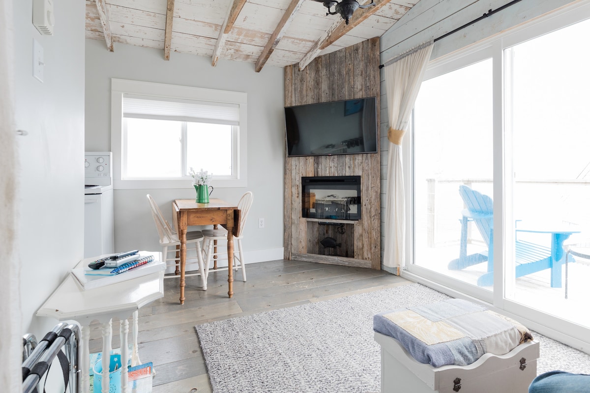 An open living area features a rustic wooden accent wall, a mounted television, and a cozy fireplace. A small dining table with two chairs is positioned near large windows, which allow ample natural light. A patterned rug covers the wooden floor, complementing the space.