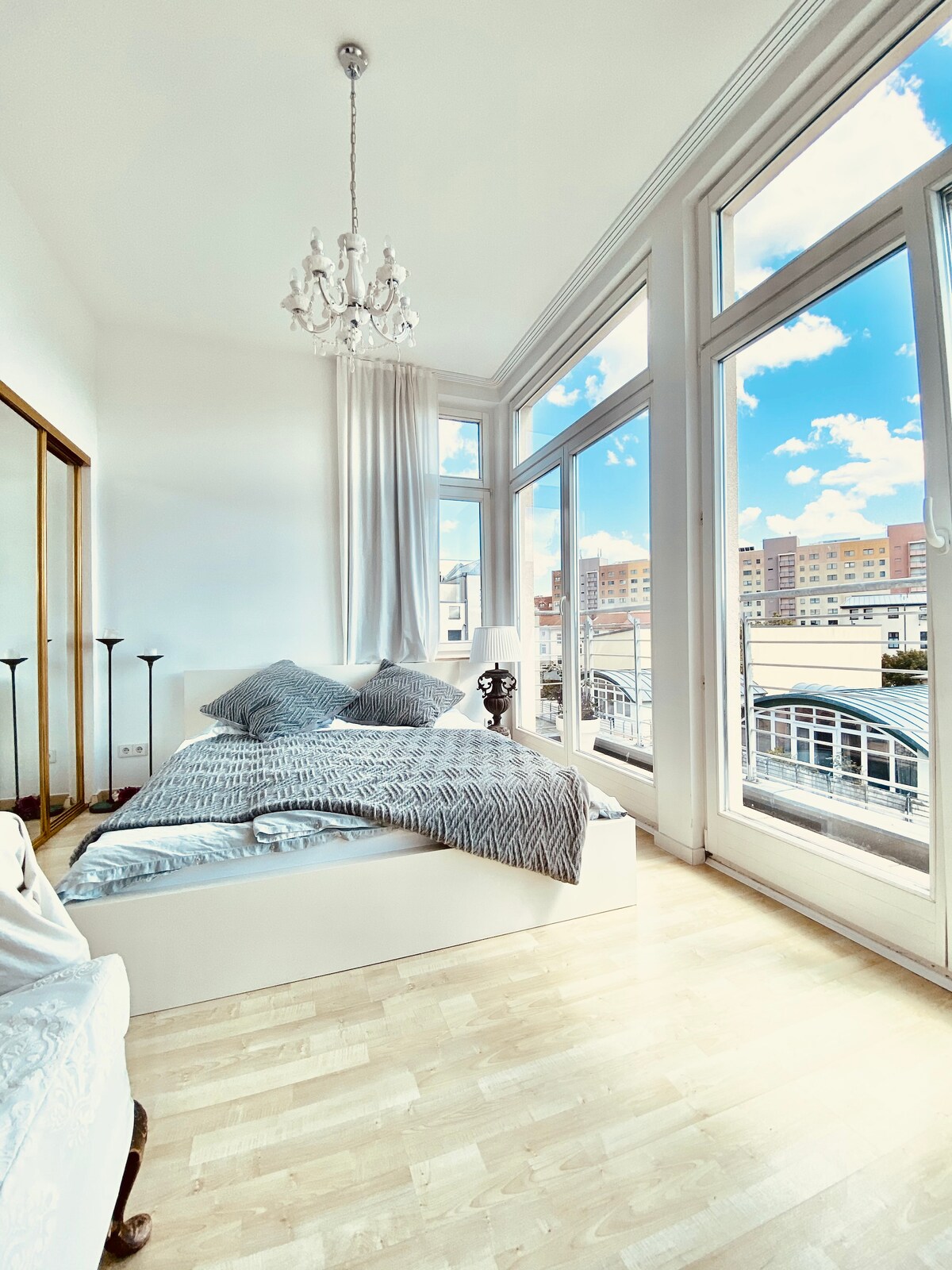 A bright bedroom features large windows allowing natural light to fill the space. A spacious bed with layered bedding is positioned against the wall, complemented by a stylish chandelier overhead. The mirrored closet adds a sense of openness and reflects the inviting atmosphere.