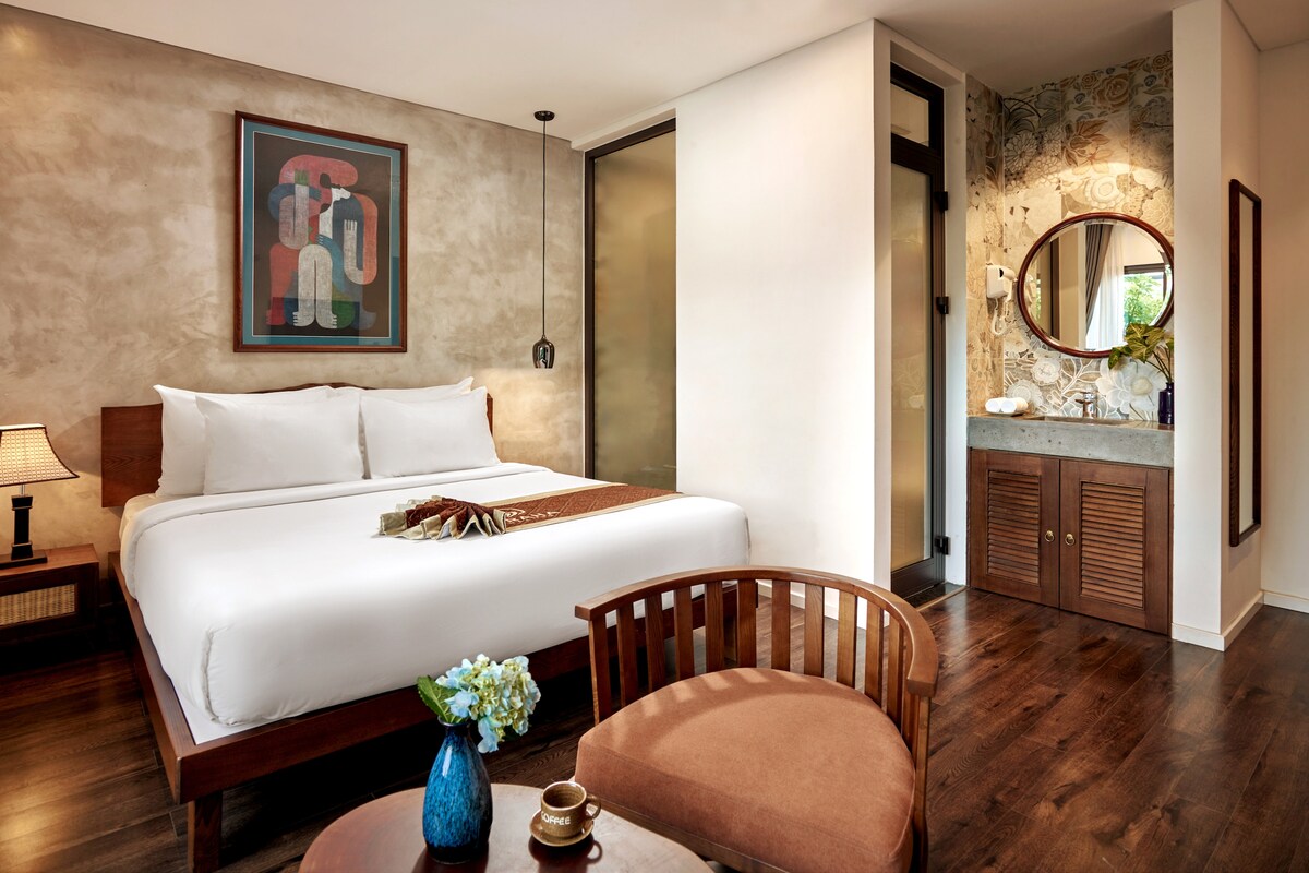 A well-appointed room features a spacious bed with crisp white linens against a textured wall. A wooden chair complements a side table with a decorative arrangement. A mirrored vanity and a bathroom area with elegant finishes are visible in the background.
