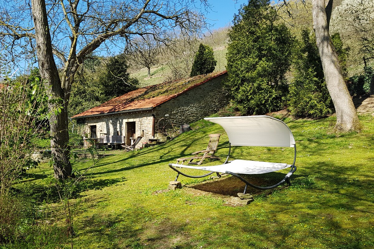 A shaded lounge area is situated on a grassy expanse, featuring a contemporary sunbed with a protective canopy. In the background, a rustic stone building with a slanted roof complements the surrounding trees and greenery, creating a serene outdoor space.