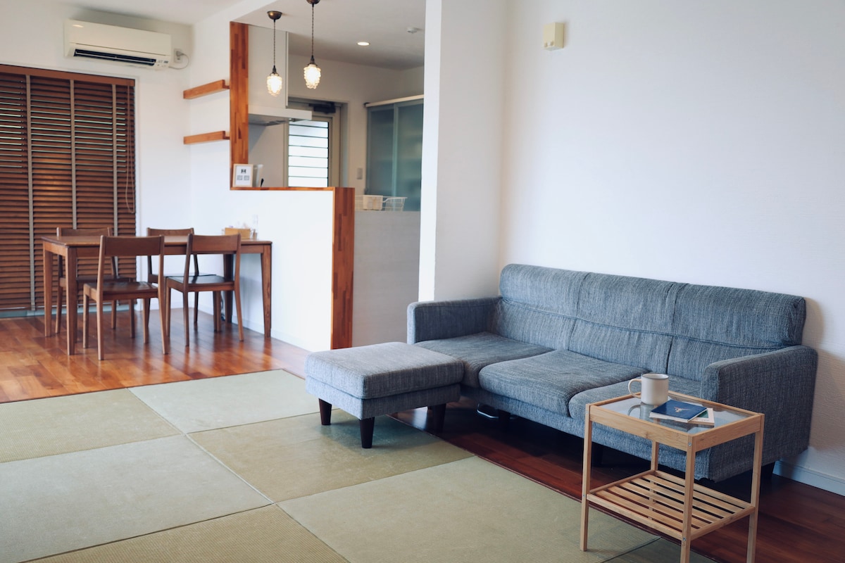 A bright living area features a cozy gray sofa with an accompanying ottoman. A wooden side table holds a cup, while light streams in from the nearby window. In the background, a dining table set for four is visible, enhancing the inviting atmosphere.