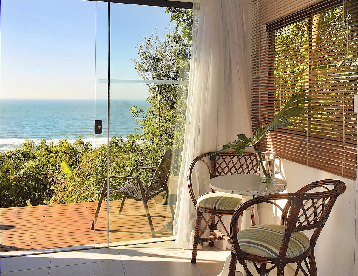 A glass door opens to a private deck with views of the ocean. A small round table and two rattan chairs are positioned nearby, surrounded by lush greenery. Natural light filters through the sheer curtains, enhancing the serene atmosphere.