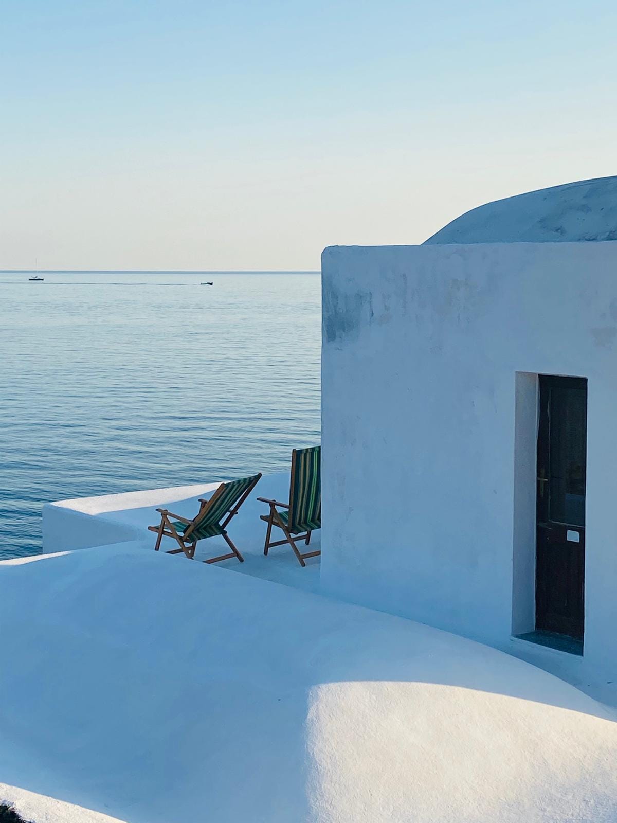 Two striped chairs are positioned on a sunlit terrace, overlooking the calm sea. The whitewashed walls reflect the soft light, while the tranquil water and distant boats contribute to a serene coastal ambiance.