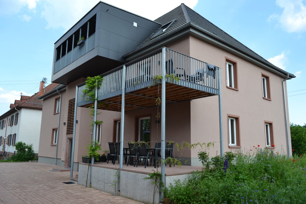 A modern two-story building features a light pink facade with a gray balcony extension above. A spacious terrace with outdoor seating is visible at the ground level. Surrounding greenery adds a natural touch to the setting.