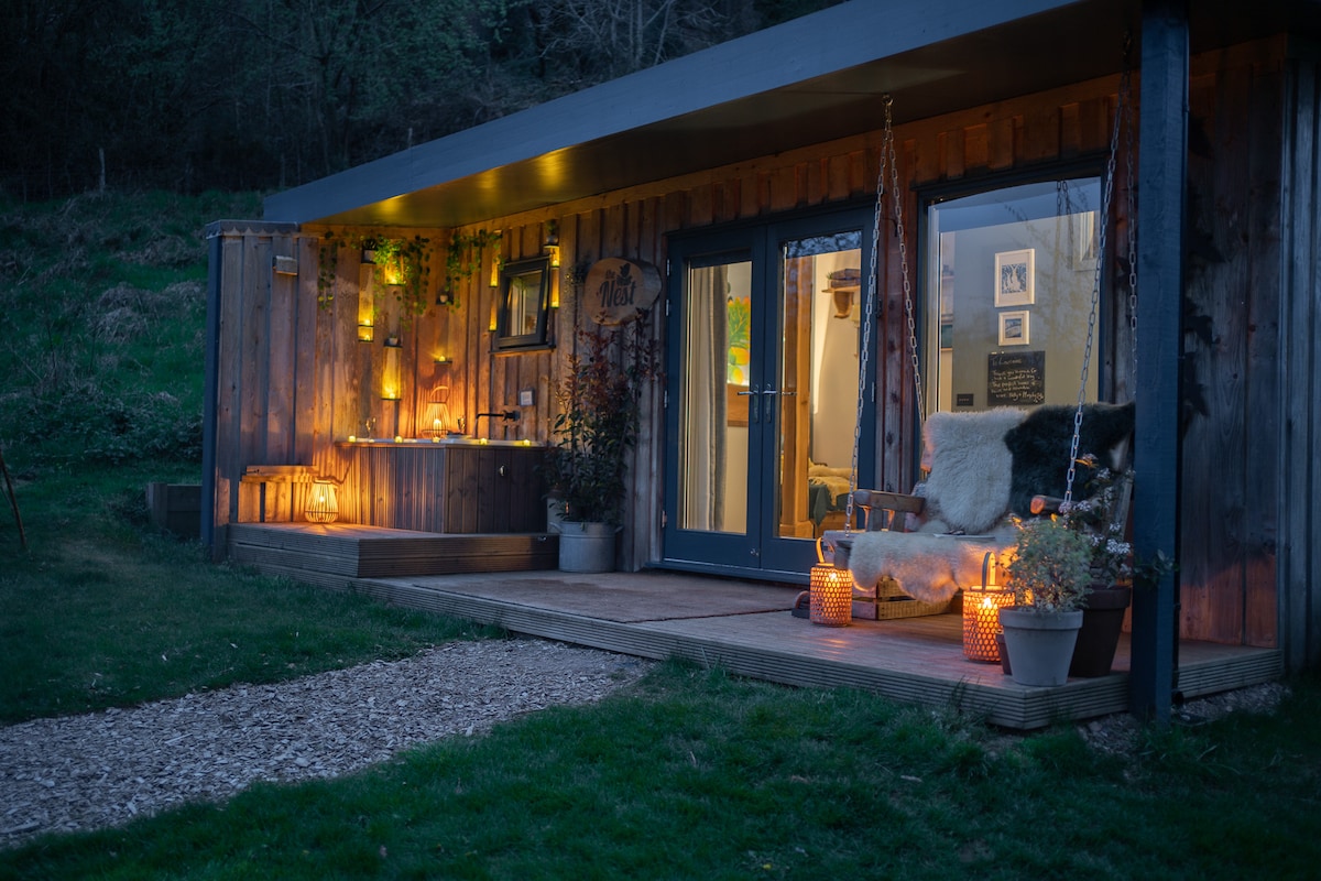 The exterior of the cabin is illuminated by soft amber lights, highlighting the wooden facade. A welcoming porch area features a cozy chair and decorative lanterns, creating an inviting atmosphere. Large glass doors provide a view into the warmly lit interior.