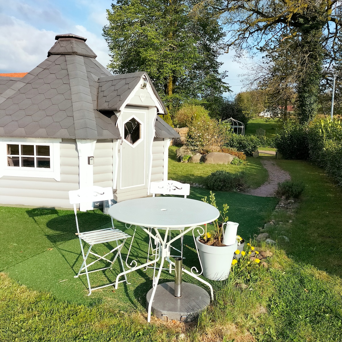 A charming cabin with a grey shingled roof is surrounded by lush greenery. A small circular table and two white chairs are positioned on a grassy area, inviting outdoor relaxation. A path leads into the garden, enhancing the serene atmosphere.
