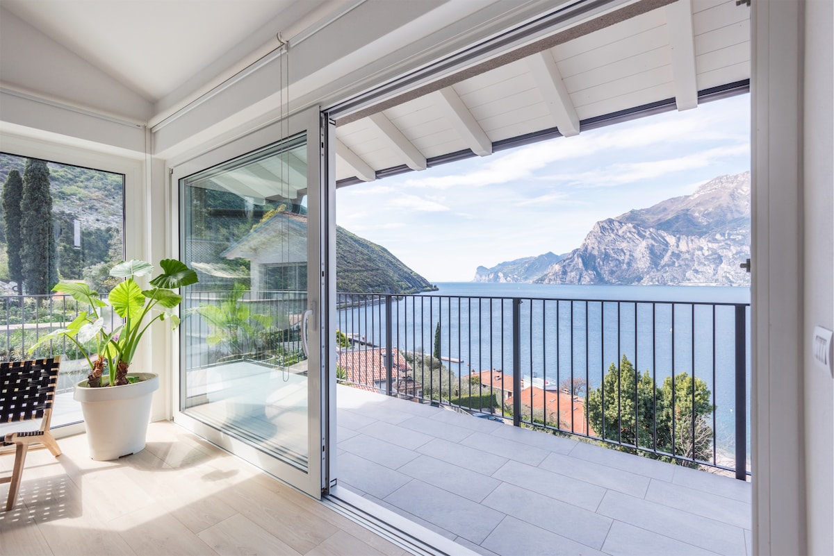 A bright living space features large glass doors that open to a balcony with views of Lake Garda and surrounding mountains. A green potted plant and minimalist woven chair are positioned beside the open doors, adding a touch of nature to the modern interior.