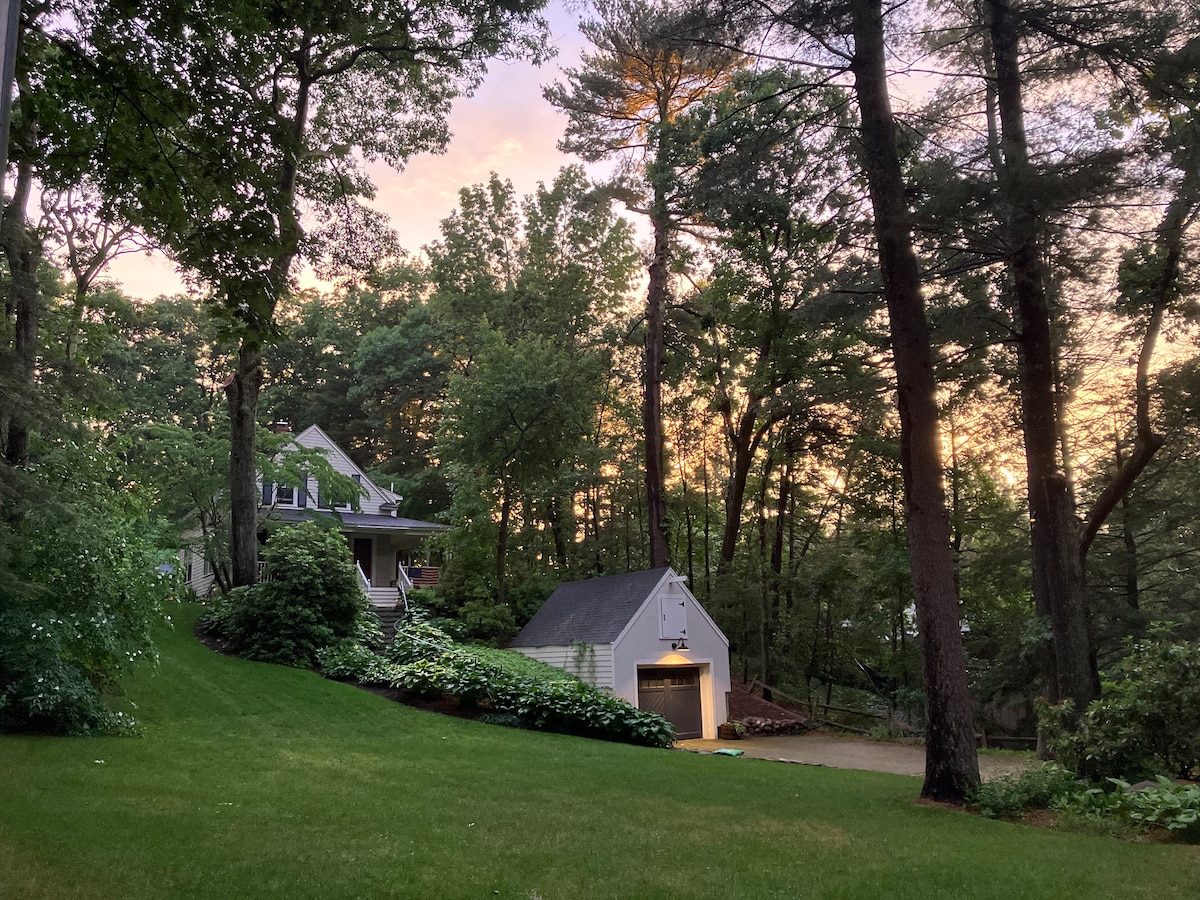 The exterior of a historic home is surrounded by tall trees, with a gentle slope of green lawn leading to the entrance. A spacious garage is visible on the right. The sky is painted in soft hues, indicating sunset.