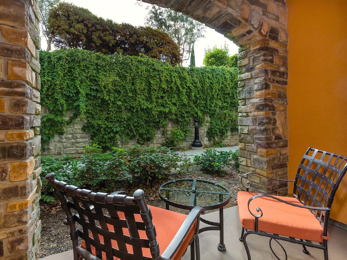 A private patio features two chairs with orange cushions and a glass-topped table, surrounded by a lush green vine-covered wall. The area is shaded by an archway made of natural stone, providing a cozy outdoor spot.