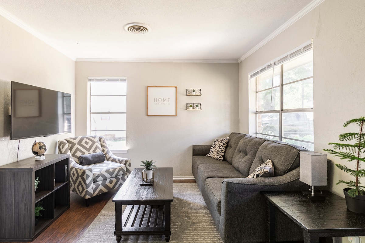 A comfortable living area features a dark gray sofa and an accent chair with a geometric pattern. A wooden coffee table is centered in the room, complemented by a small plant. Natural light streams in through two windows, illuminating the space.