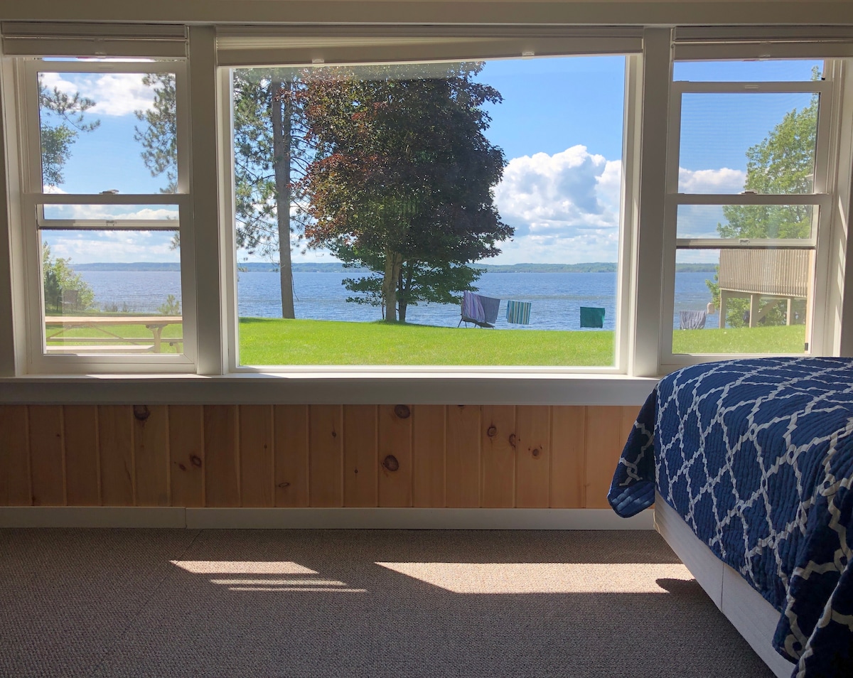 A serene bedroom offers expansive windows with views of Lake Michigan and a lush green yard. A cozy bed with a patterned blanket sits adjacent to natural light pouring in, enhancing the room's spacious ambiance. Outside, trees and a picnic table are visible.