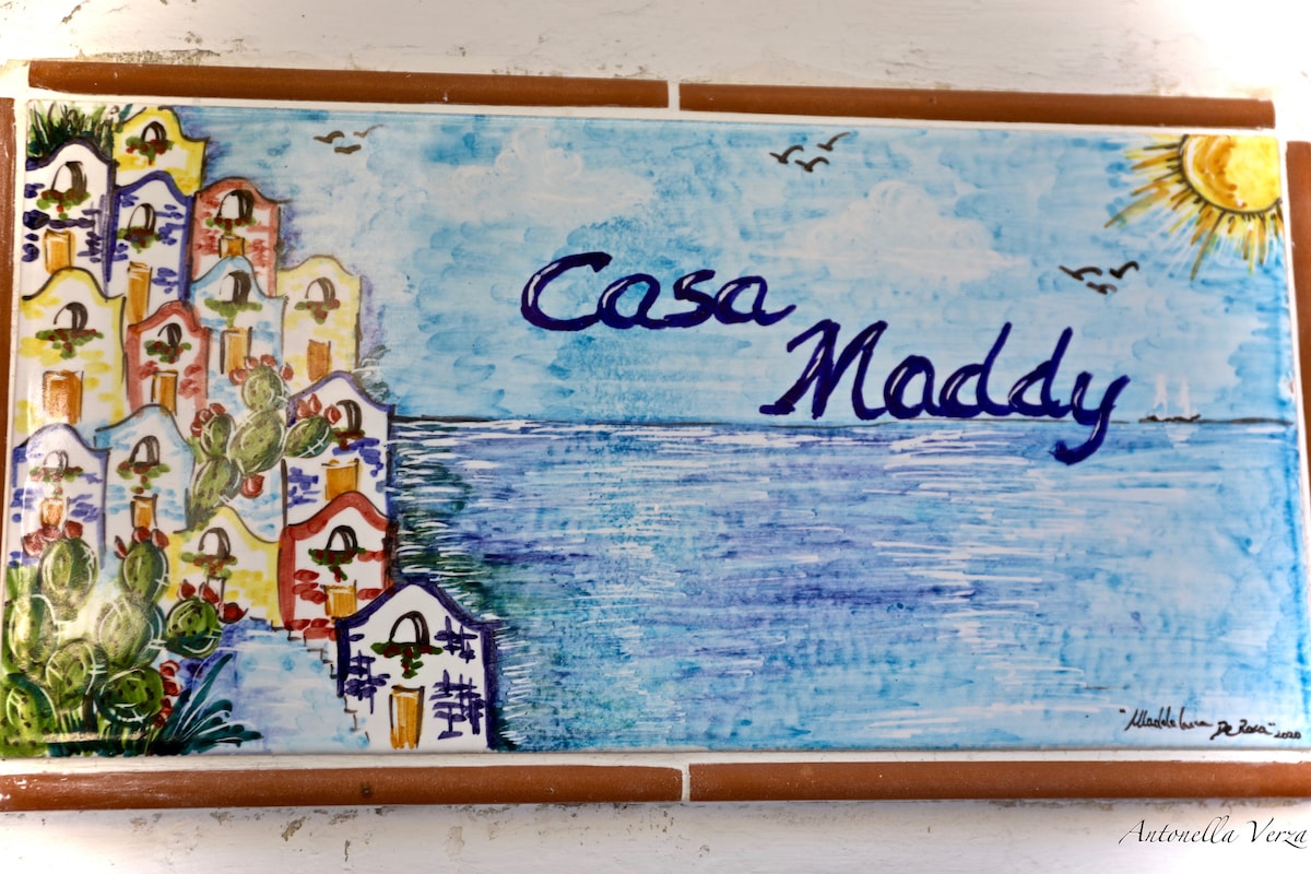 A hand-painted tile sign displays the name 'Casa Maddy' in a flowing blue script. The artwork features a coastal scene with colorful houses alongside the sea, framed by a sun and seagulls in a bright sky.