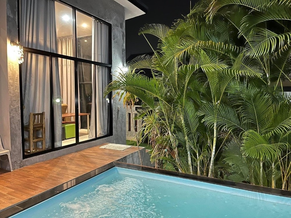 A private swimming pool is framed by lush greenery, with the exterior of a modern villa visible in the background. Large glass windows illuminate the indoor seating area with soft lighting, blending indoor and outdoor elements harmoniously.