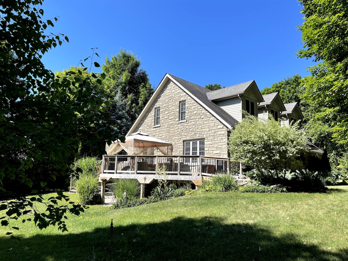 A charming stone house is set amidst lush greenery, highlighting a spacious deck with a canopy. Sunlight illuminates the well-maintained landscaping, which features various plants and trees around the property, presenting an inviting outdoor space.