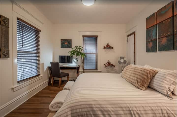 The bedroom features a king-size bed with white linens, 4 king-size down pillows & a body pillow - all of which rests on top of a modern pallet bed frame. The bedroom also has a place to hang clothes, a mirror, and extra blankets. 