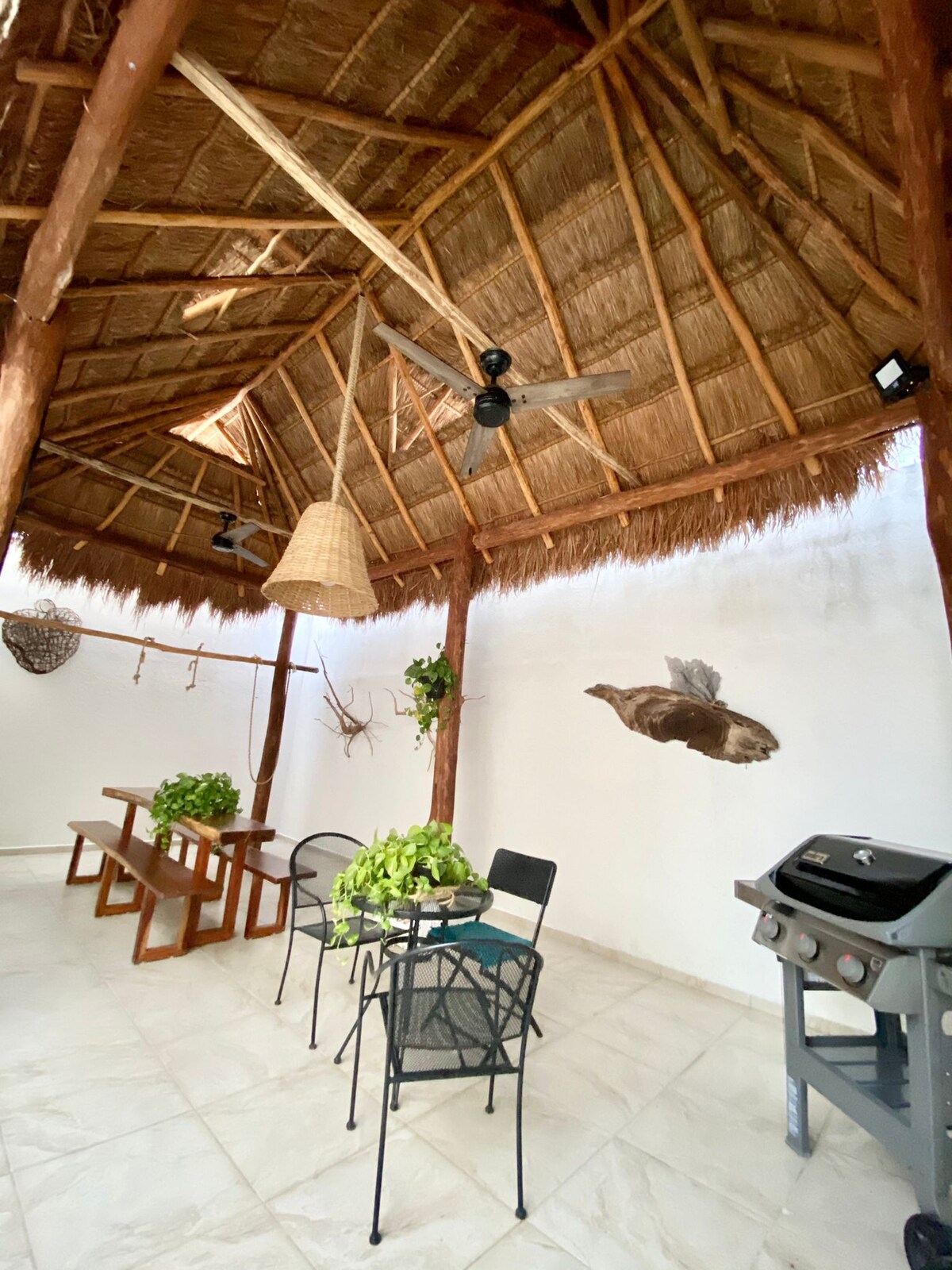An outdoor space with a thatched roof showcases a long wooden table surrounded by black chairs. A grill is positioned at one side, while greenery decorates the area. Natural light filters through, creating a relaxed environment that encourages gathering.