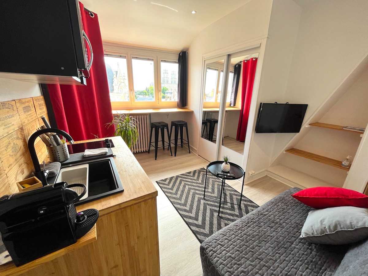 A compact studio features large windows dressed in red curtains, allowing natural light to fill the space. A kitchenette with wooden countertops is visible alongside a small dining area with high stools. A cozy seating area is arranged with a convertible sofa and a decorative rug.