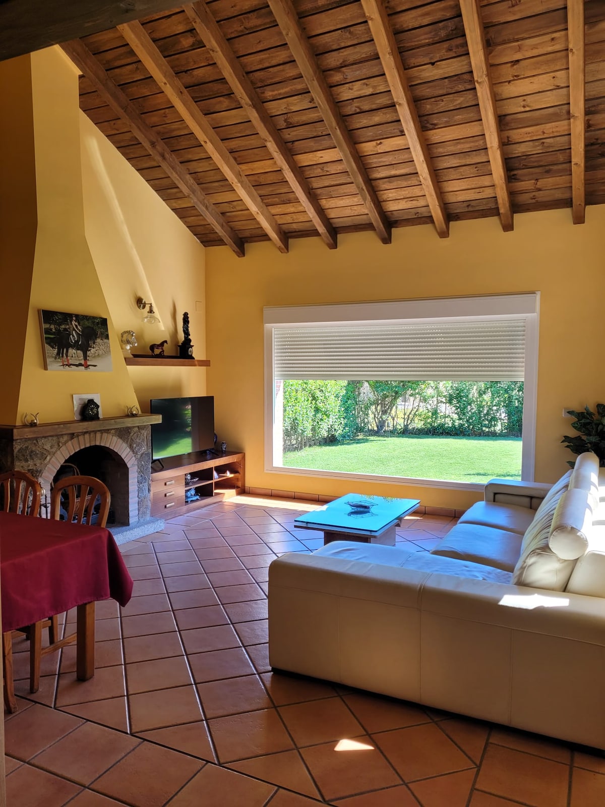 An airy living space features a high, wooden ceiling and a large window that allows natural light to fill the room. A comfortable white sofa faces a wooden coffee table, with an entertainment unit positioned nearby. A fireplace adds a cozy element to the area.