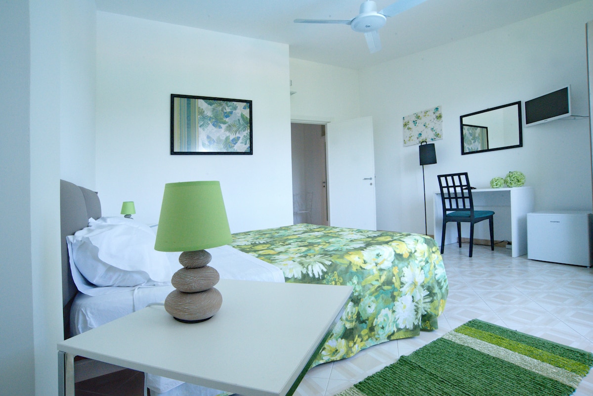 A bright and spacious room features a large bed with floral bedding, complemented by a ceiling fan. A simple wooden side table with a green lamp is positioned beside the bed. A small desk and chair are visible, along with decorative wall art that enhances the light atmosphere.
