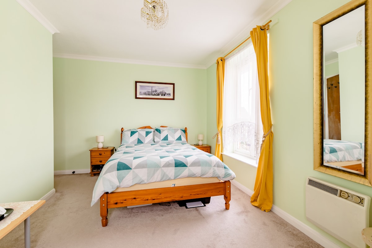 A spacious double bedroom features soft green walls and natural light streaming in through a large window dressed with yellow curtains. A wooden bed with a geometric patterned duvet cover is complemented by two bedside tables with lamps. A decorative mirror adds depth to the room.