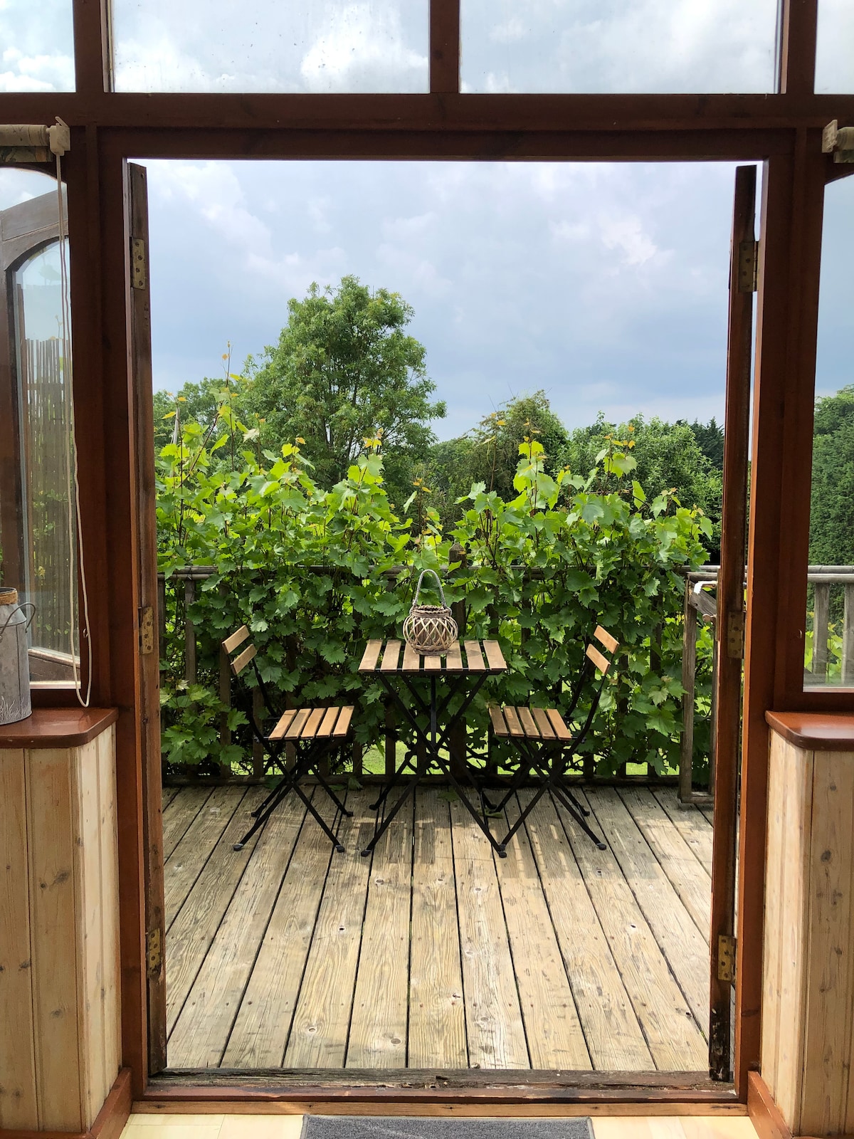 A welcoming terrace is displayed, featuring a wooden deck surrounded by lush greenery. A small table and two chairs offer an inviting space for relaxation. The open doors create a seamless connection between indoor and outdoor areas, allowing natural light to fill the room.
