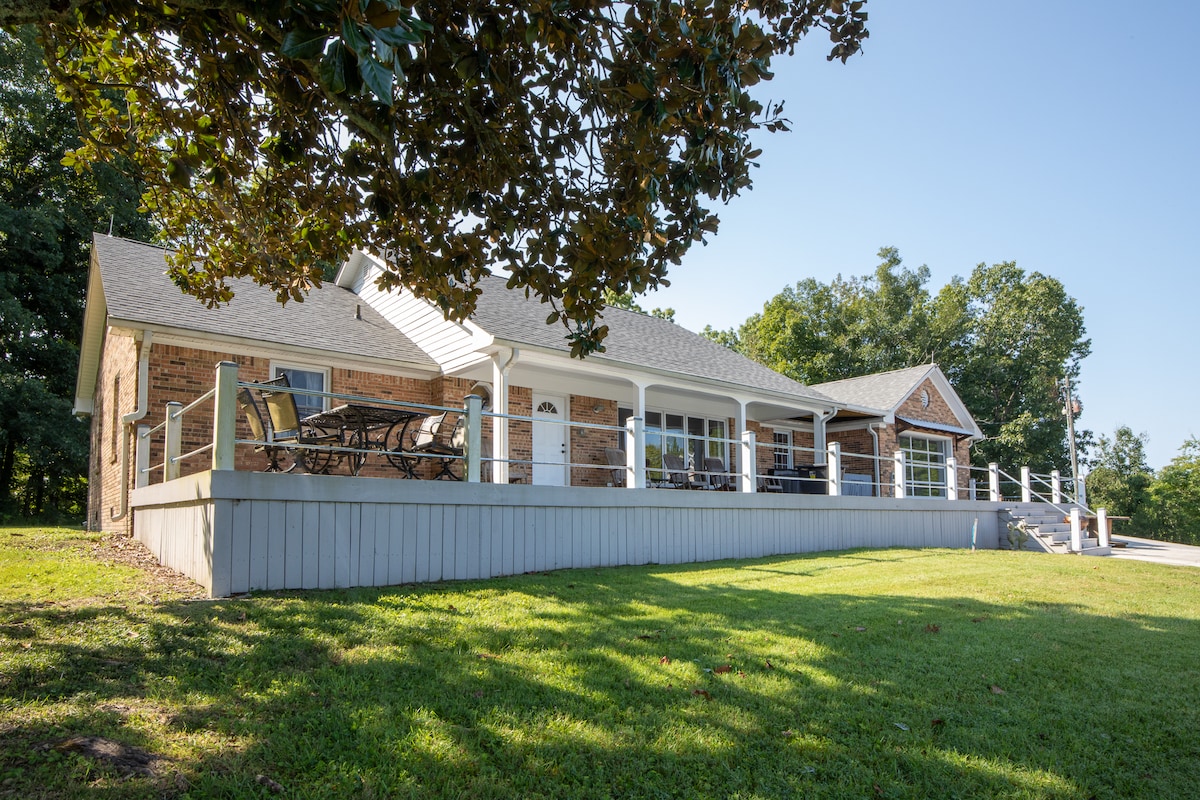 A hilltop home features a spacious patio with outdoor seating, framed by lush greenery. The brick facade is complemented by a well-maintained lawn, providing ample space for relaxation. The roofline and porch create a welcoming entrance, inviting guests to enjoy the serene surroundings.