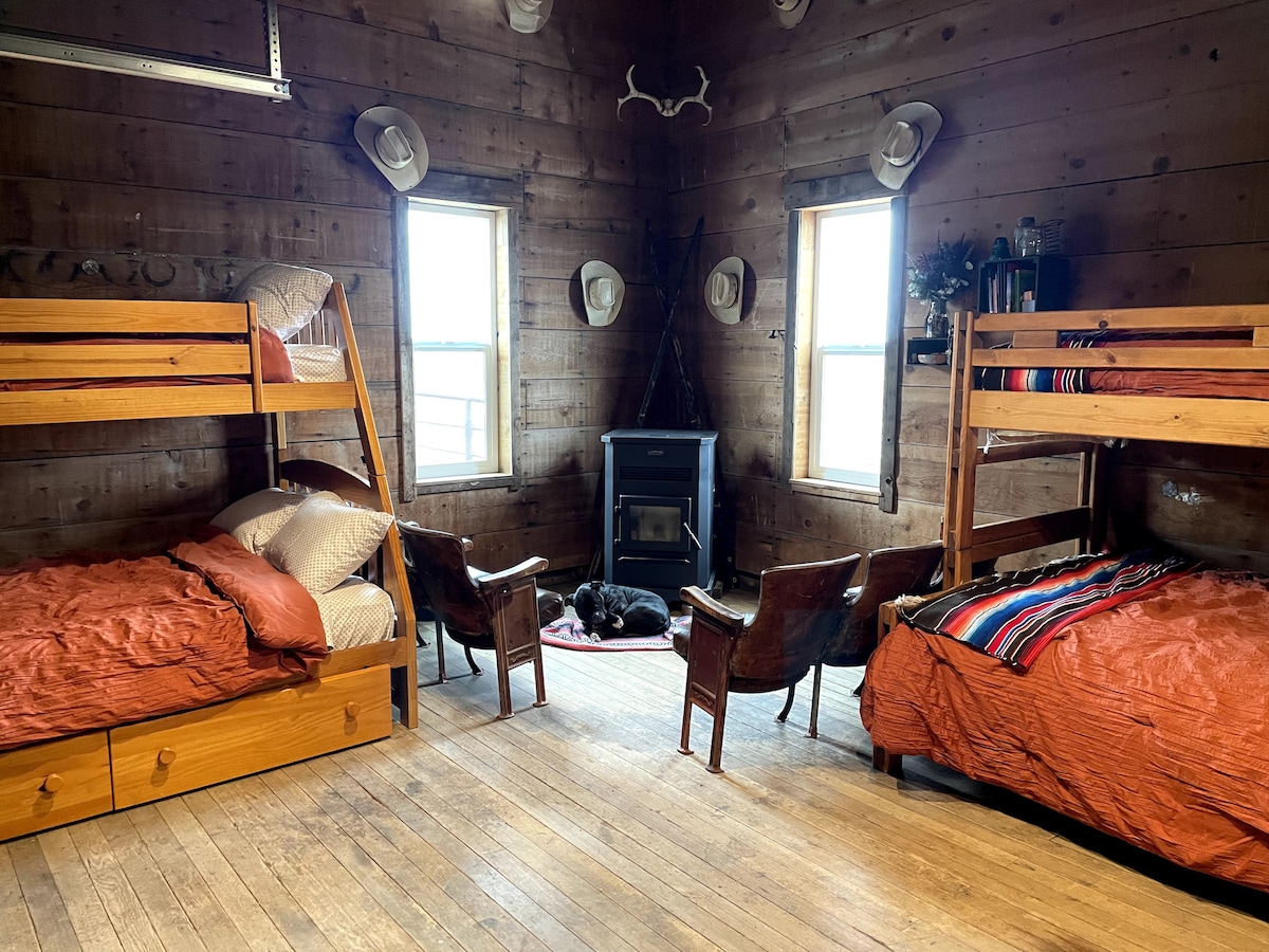 An open concept sleeping area features two sets of wooden twin over queen bunk beds, dressed in warm-toned linens. Two vintage chairs are situated across from a compact stove, with a soft blanket on the floor. Natural light is welcomed through two windows framed by rustic wood walls.